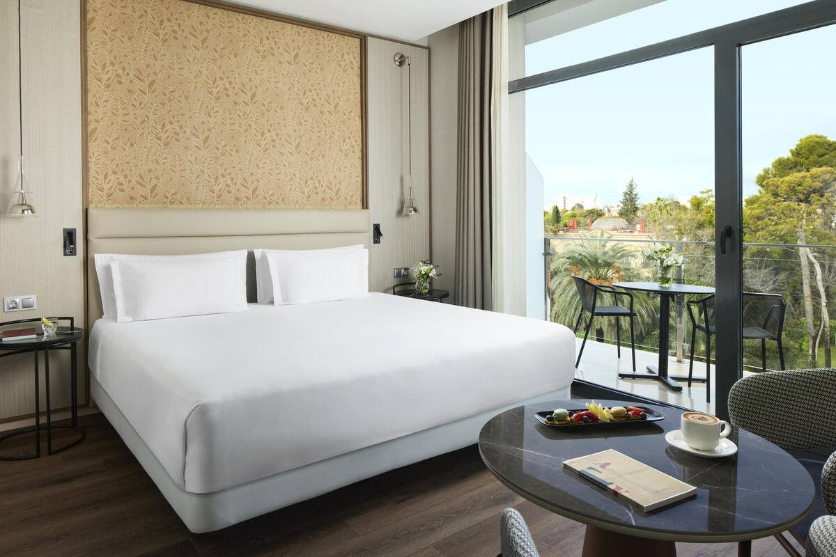 A hotel room with a large bed, a balcony with a table and chairs, and a view of the outdoors.