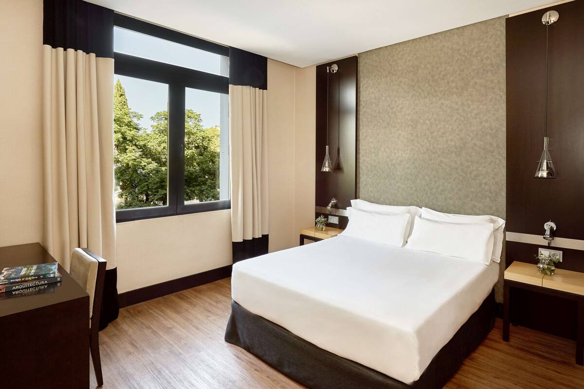 A hotel room with a large bed, a desk with a lamp, a nightstand, and a view of trees through the window.