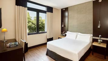 Habitación de hotel con una cama grande, un escritorio con lámpara, una mesita de noche y vista a árboles desde la ventana.