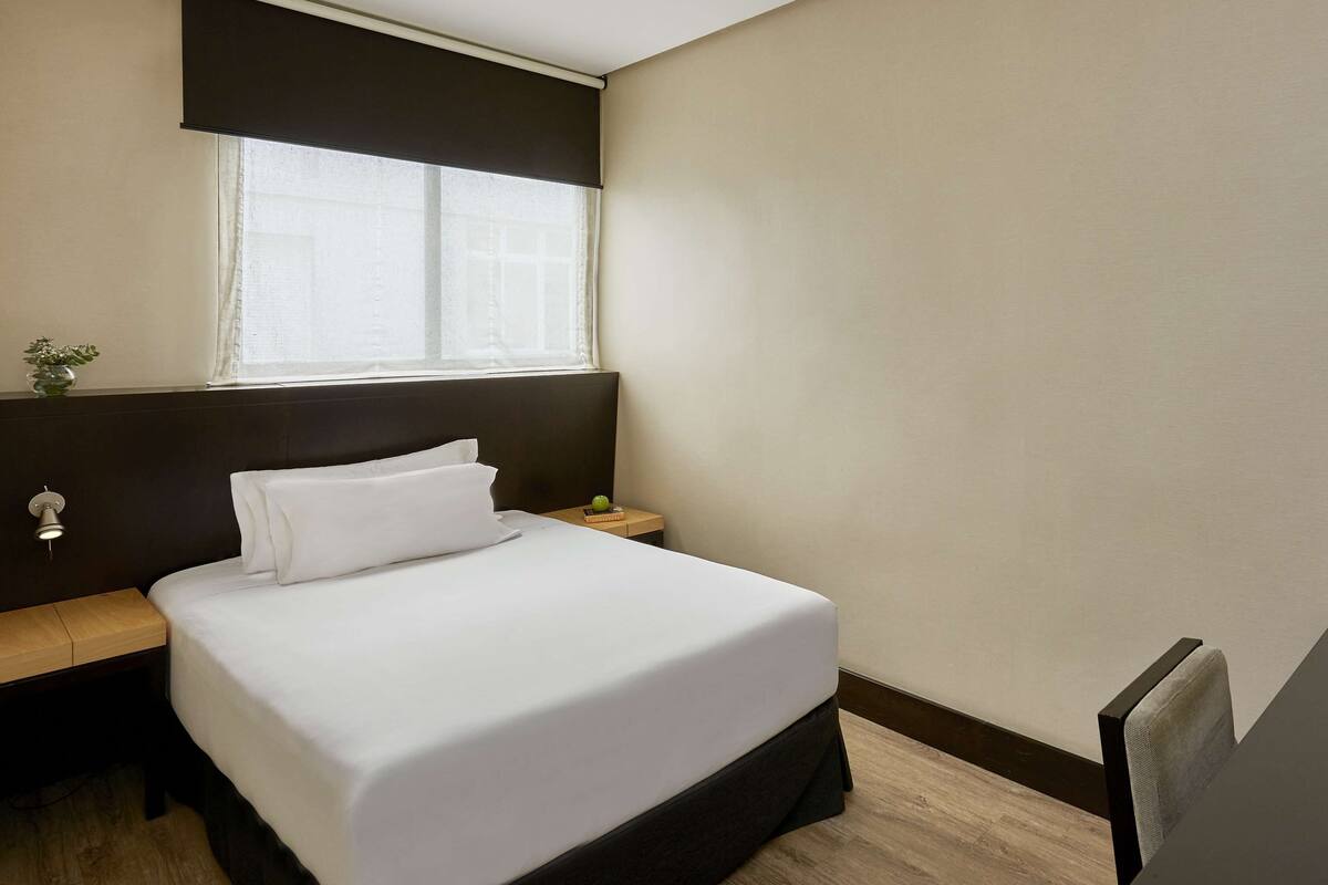 A hotel room with a bed, bedside table, lamp, and a view of the outside.
