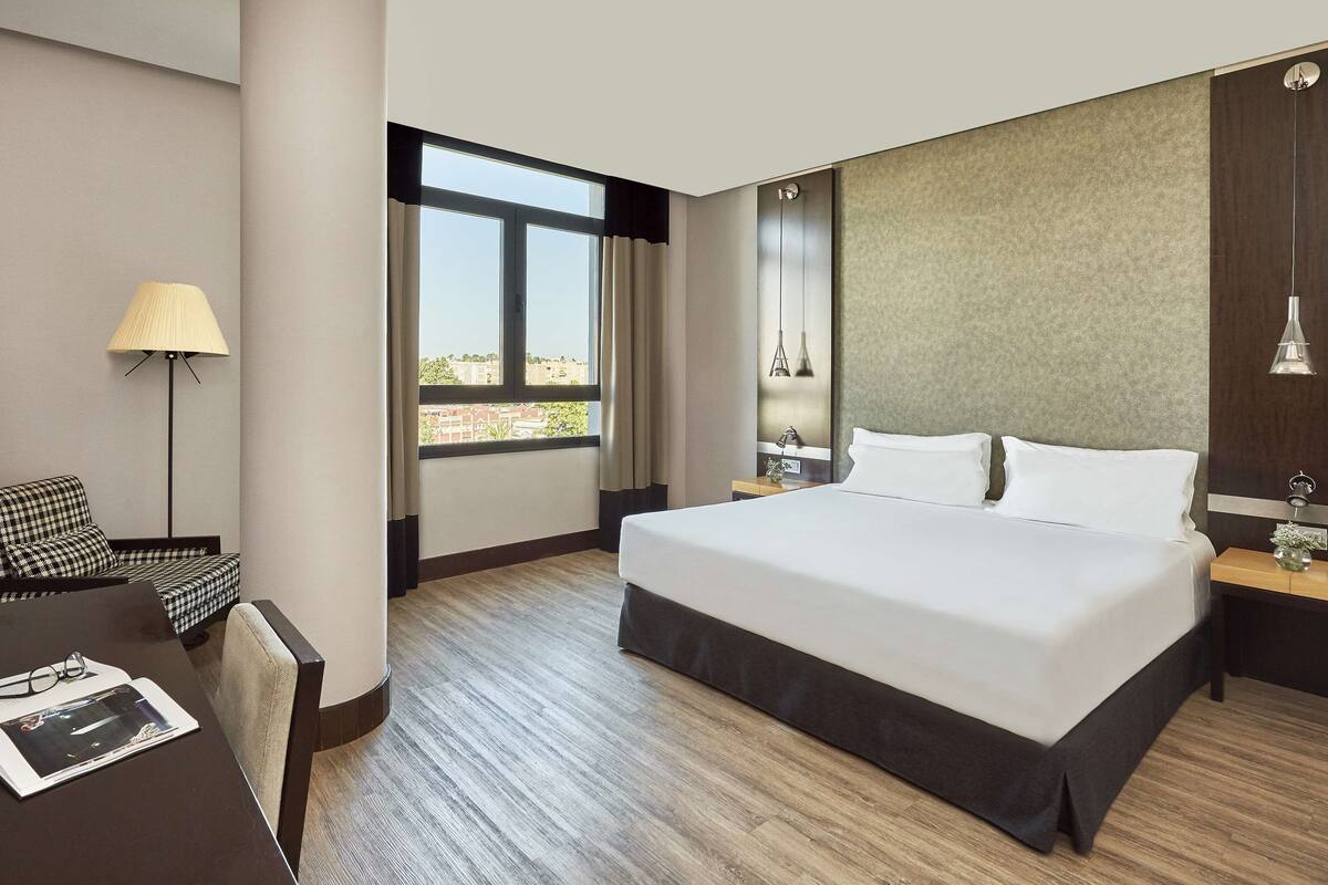 A hotel room with a large bed, a desk with a lamp, a chair, and a view of the city through the window.