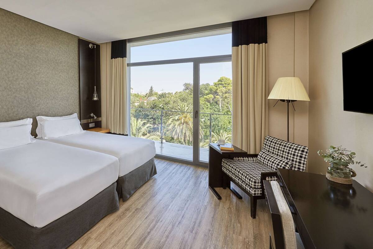 A hotel room with two beds, a desk, a chair, a TV, and a balcony with a view of trees.