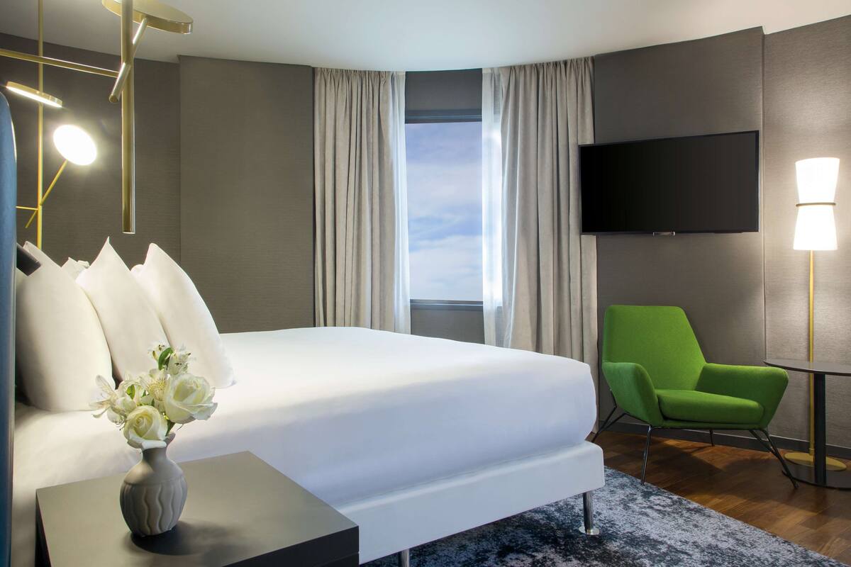 A hotel room with a bed, a green armchair, a flat-screen TV, and a vase with flowers.