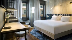 Une chambre d’hôtel moderne, dotée d’un grand lit, d’un bureau, d’une chaise et offrant une vue sur un immeuble par la fenêtre.