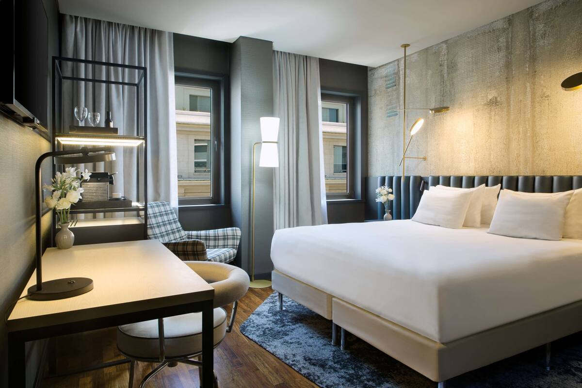 A modern hotel room with a large bed, a desk, a chair, and a view of a building through the window.