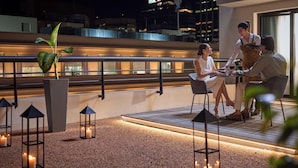 Un espace de restauration aménagé sur le toit, offrant une vue sur les gratte-ciel de la ville, notamment sur un immeuble de la HSBC. On y trouve des tables et des chaises, et un serveur est présent.