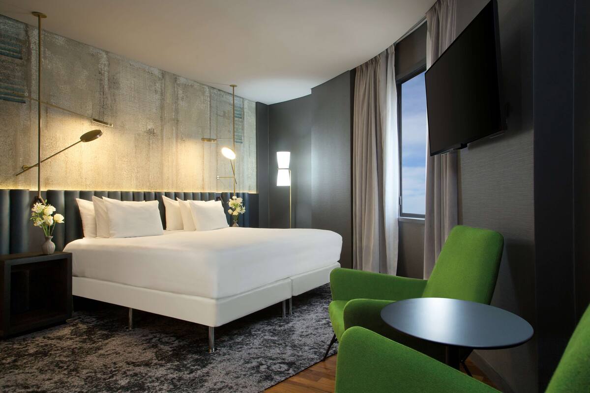 A modern hotel room with a large bed, a green armchair, a small table, and a TV mounted on the wall.