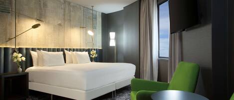 A modern hotel room with a large bed, a green armchair, a small table, and a TV mounted on the wall.