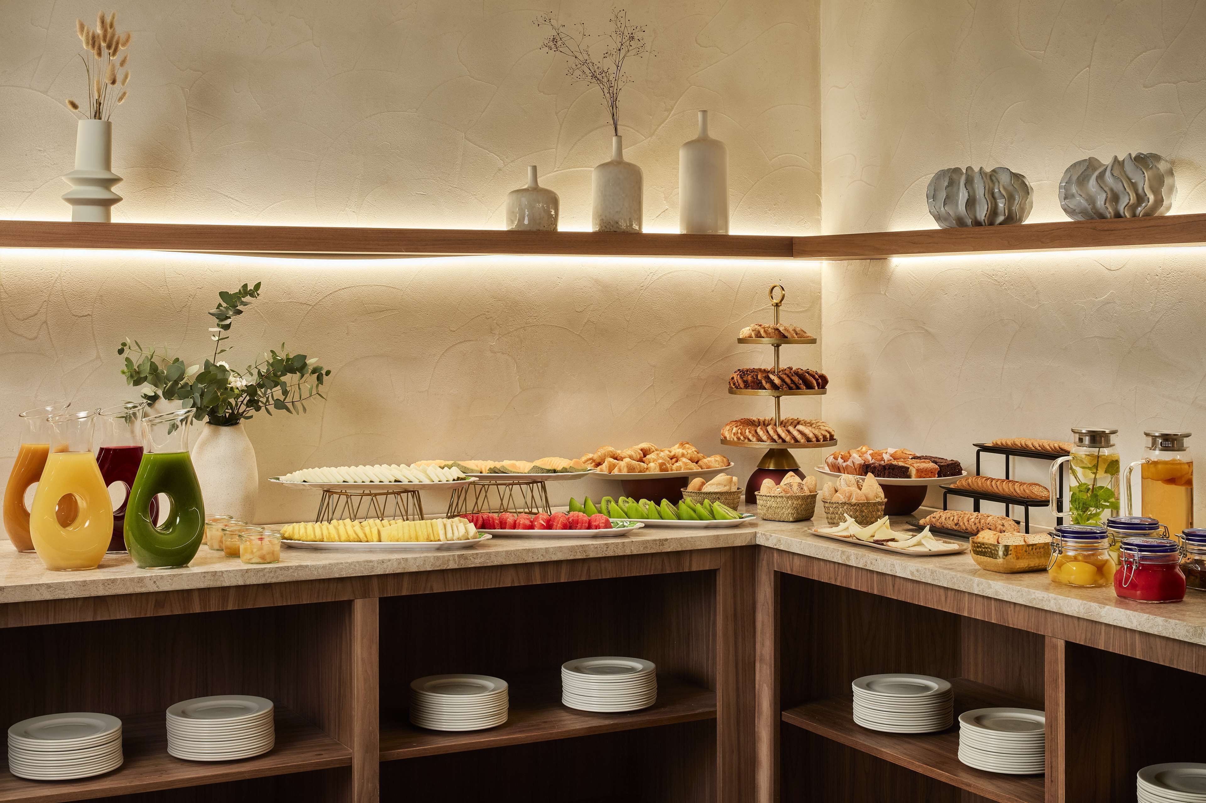 A buffet with various food items and drinks, including fruits, pastries, and beverages, arranged on a counter with shelves above.