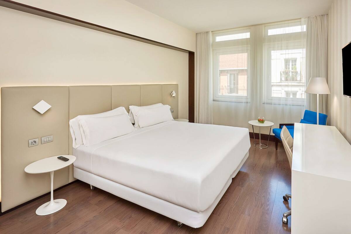 A hotel room with a large bed, a small round table, a chair, a TV, and a window with curtains.