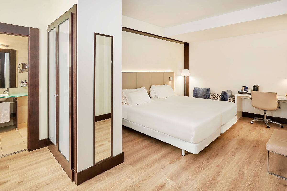 A hotel room with a large bed, a desk, a chair, a mirror, and a bathroom visible through a glass door.