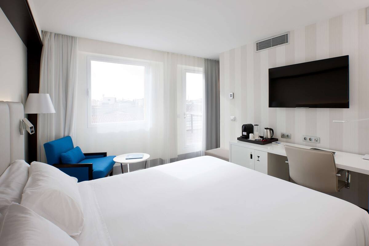 A hotel room with a large bed, a desk, a chair, a small table, a TV, and a window with curtains.