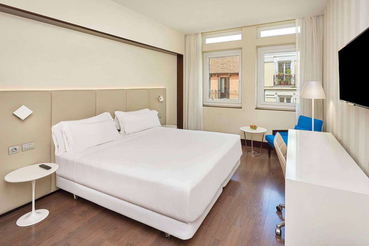 A hotel room with a large bed, a small round table, a TV, and a window with a view of buildings.