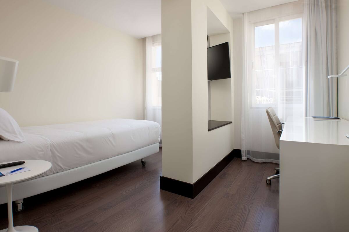 A hotel room with a single bed, a desk, a chair, a TV mounted on the wall, and a window with sheer curtains.