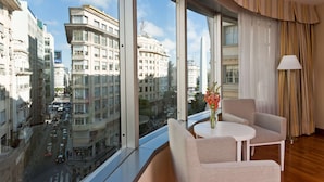 Une chambre d’hôtel dotée de grandes fenêtres donnant sur la ville, une table ronde avec deux chaises et un vase rempli de fleurs.
