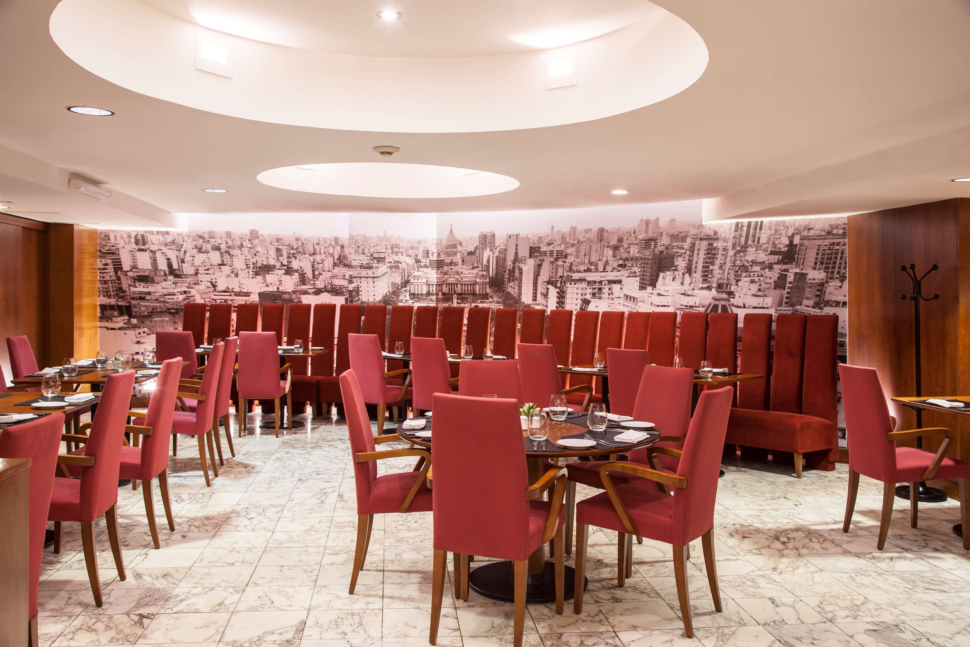 Un restaurante con sillas y mesas rojas, un mural con vista a la ciudad en la pared y un diseño de techo circular.