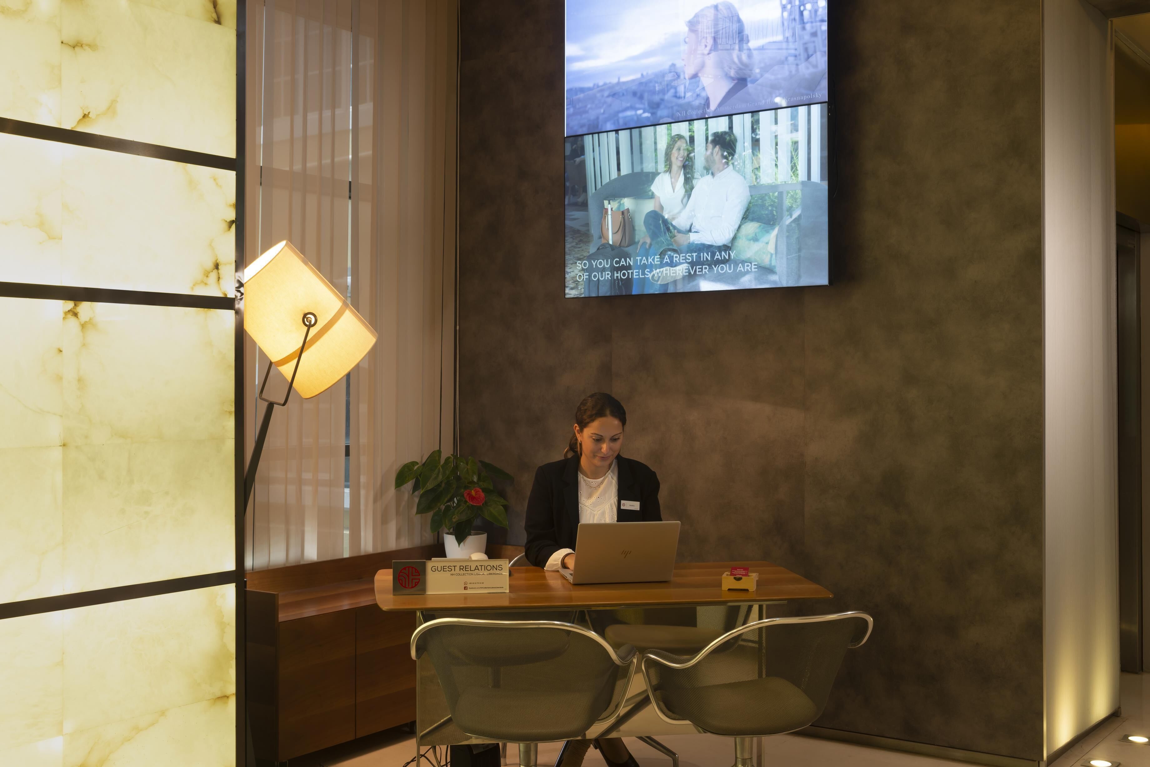 A reception desk with a laptop, a guest relations sign, and a TV screen displaying a hotel scene.
