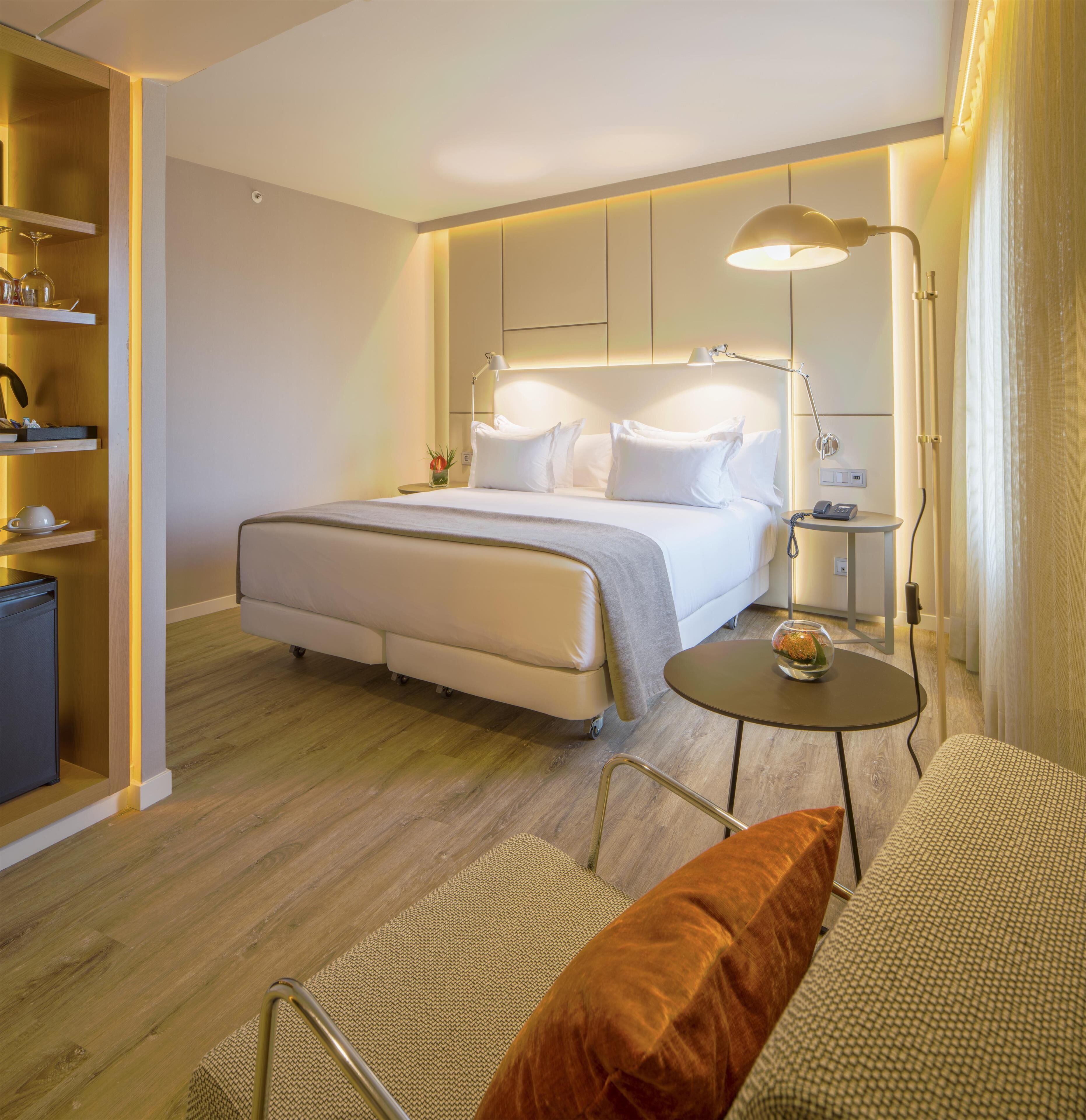 A modern hotel room with a bed, bedside table, lamp, and a small round table with a vase.
