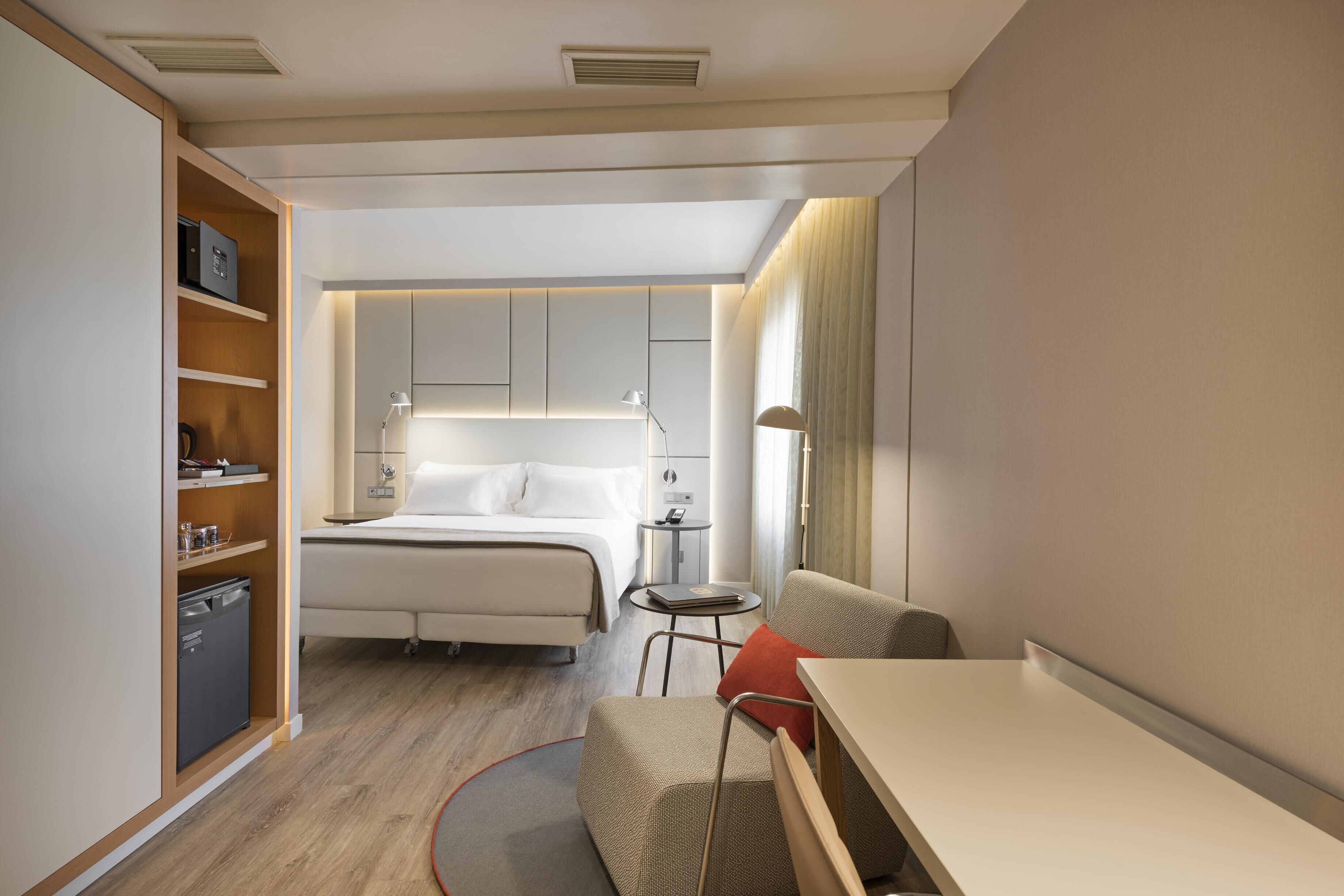 A compact hotel room with a bed, bedside table, chair, desk, and built-in wardrobe.