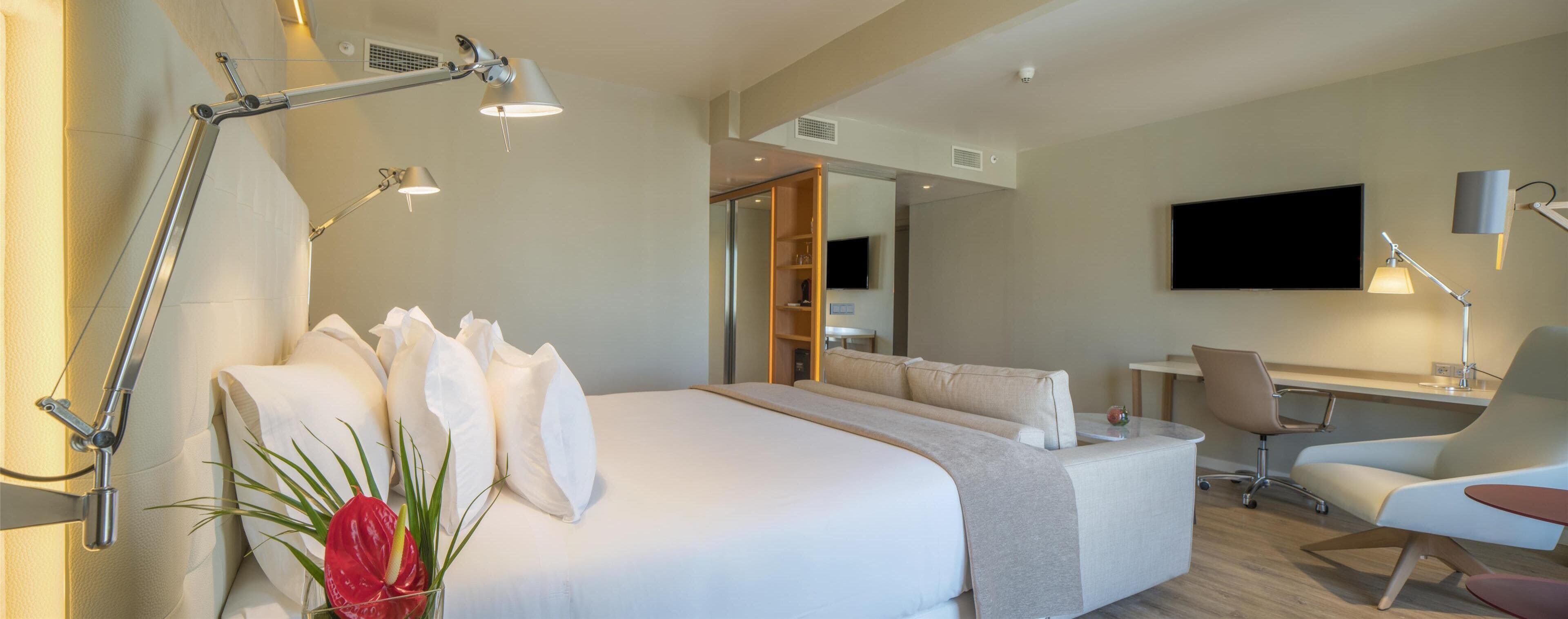 A modern hotel room with a large bed, bedside lamps, a desk with a chair, a flat-screen TV, and a plant.