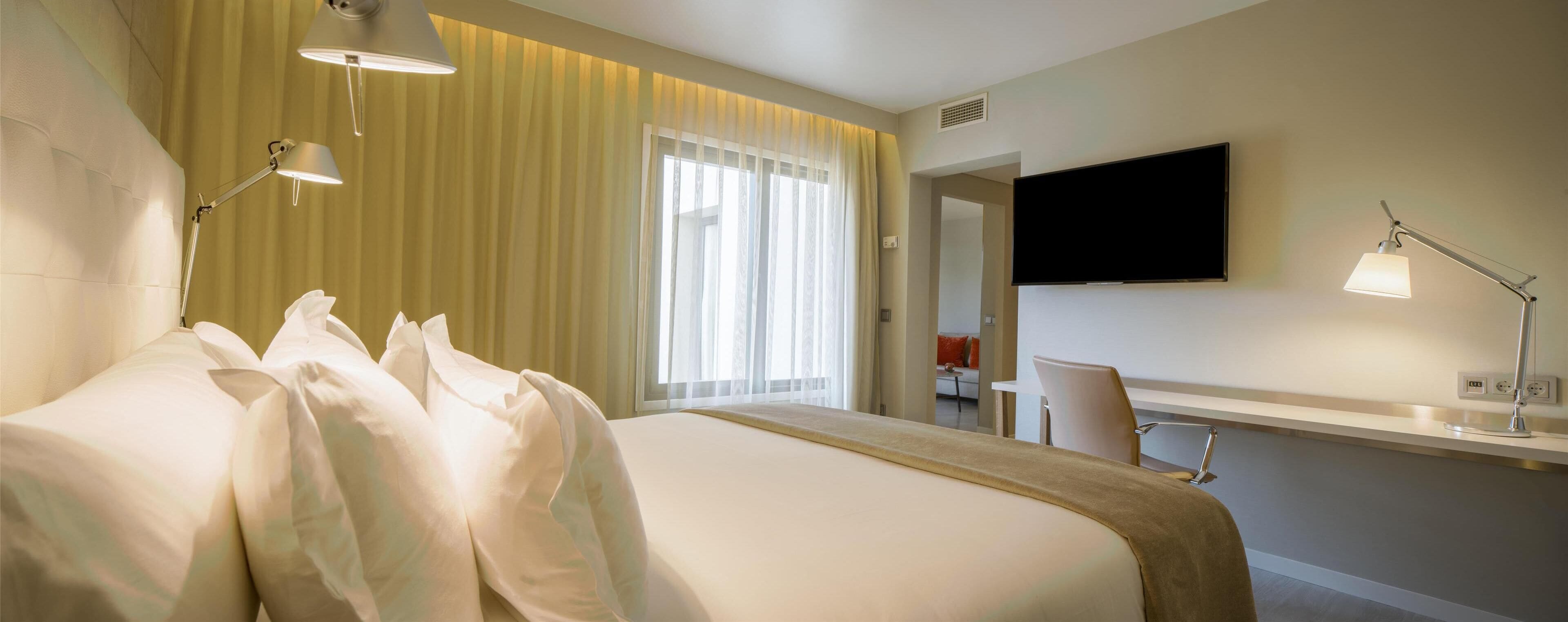 A hotel room with a bed, bedside lamps, a TV, a desk, and a chair.