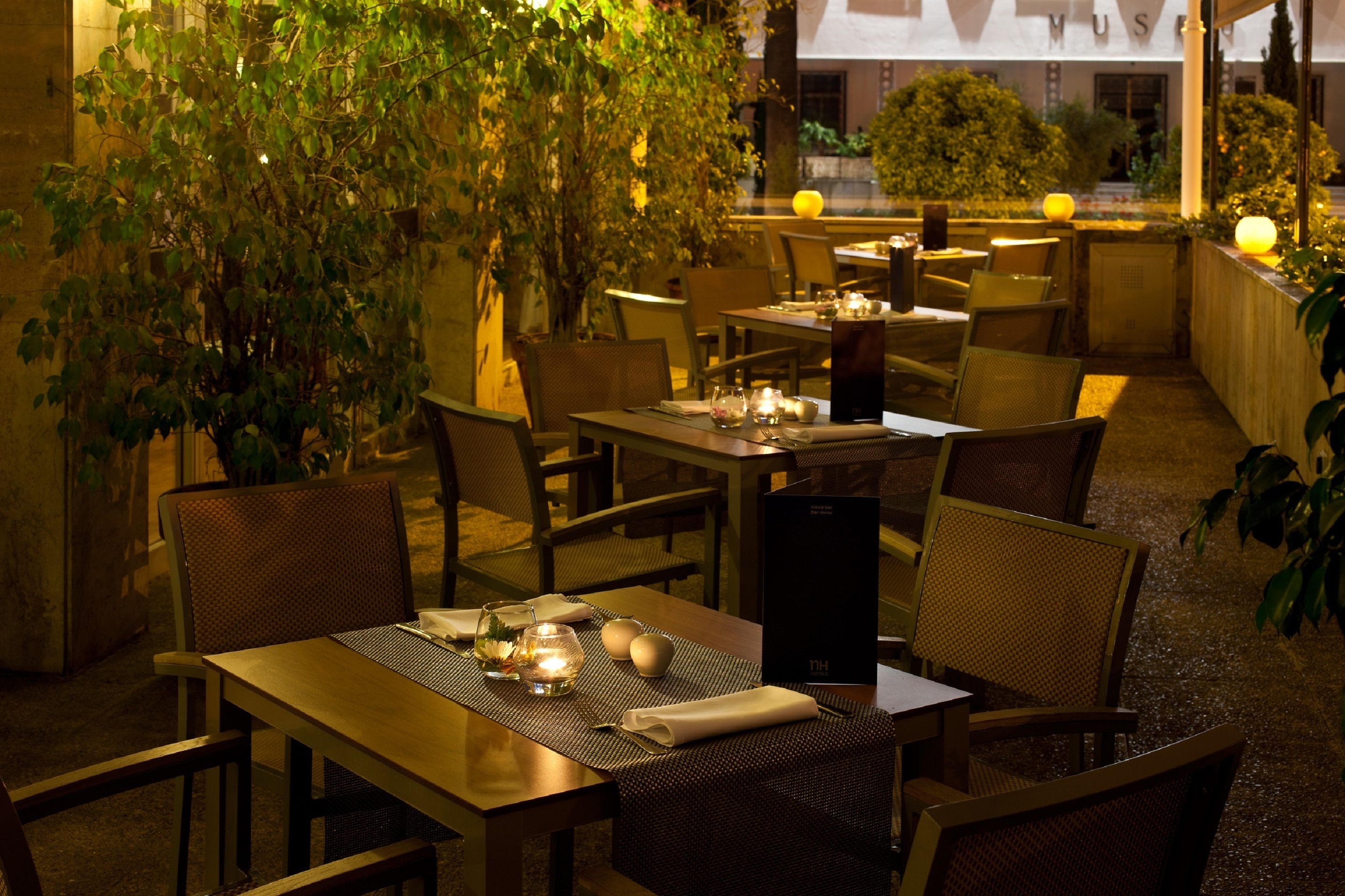 Un área de comedor al aire libre con mesas preparadas para dos, rodeada de sillas y plantas en macetas.