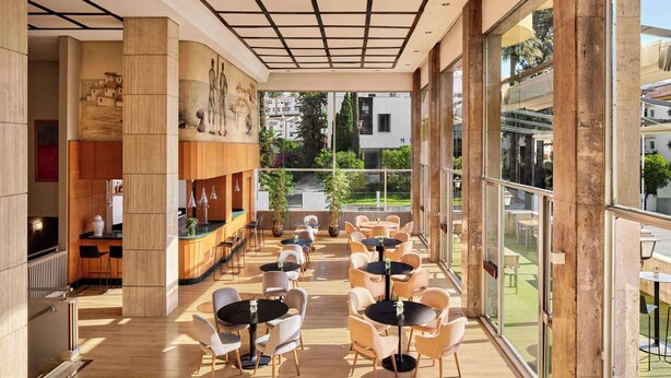 A spacious indoor area with multiple tables and chairs, large windows offering views of the outside, and a modern architectural design with a grid ceiling. - NH Luz Huelva (Huelva)