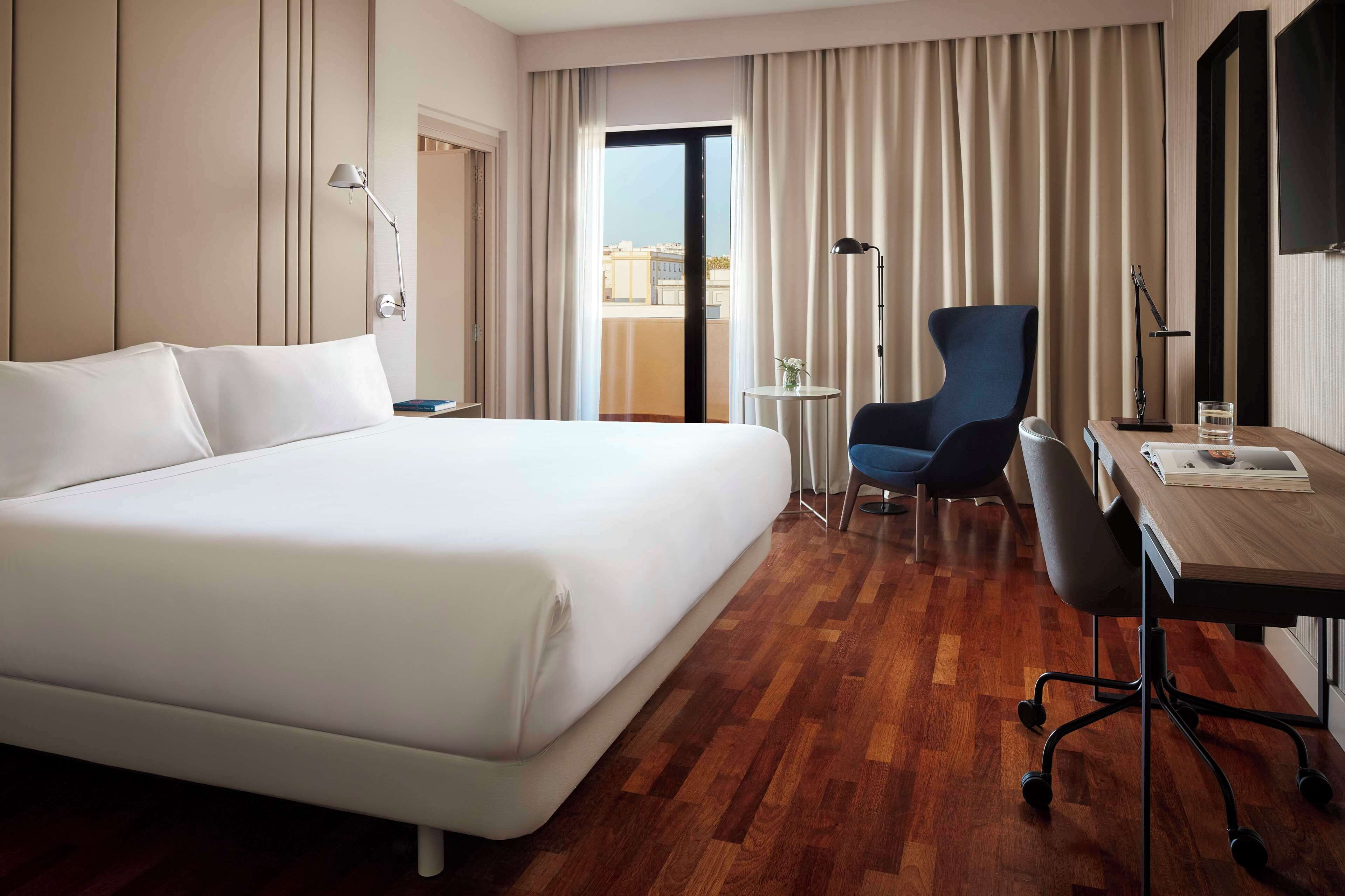 Una habitación de hotel con una cama grande, un escritorio con silla, una mesita y vistas a través de una ventana con cortinas.
