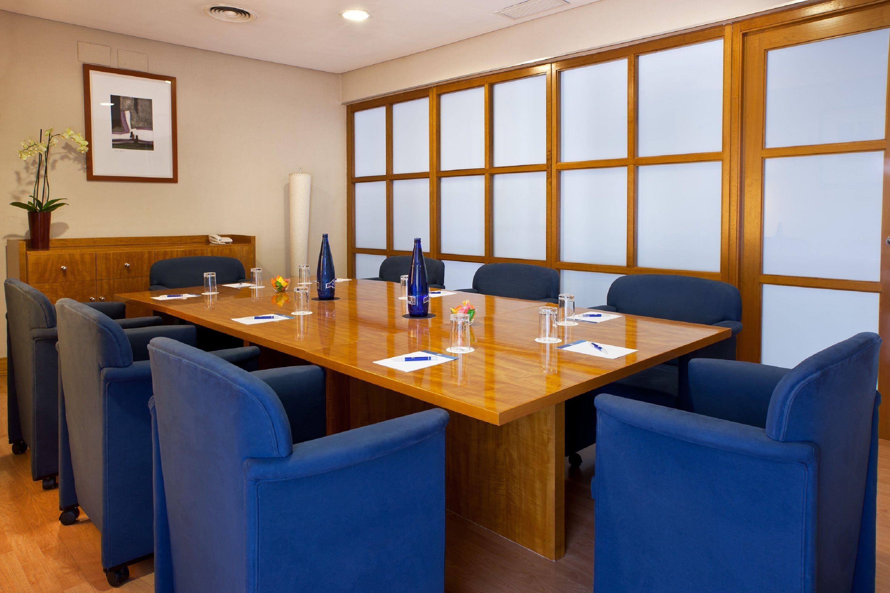 Un salón de conferencias con una larga mesa de madera, sillas azules y un amplio ventanal con paneles de vidrio esmerilado.
