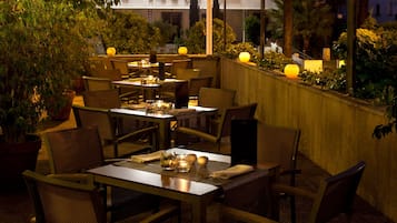 An outdoor dining area with tables and chairs, surrounded by plants and illuminated by soft lighting.