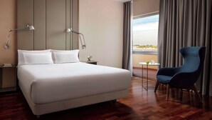 A modern hotel room with a large bed, two bedside lamps, a blue armchair, a small table with a glass, and a view of the outdoors through large windows. - NH Luz Huelva (Huelva)