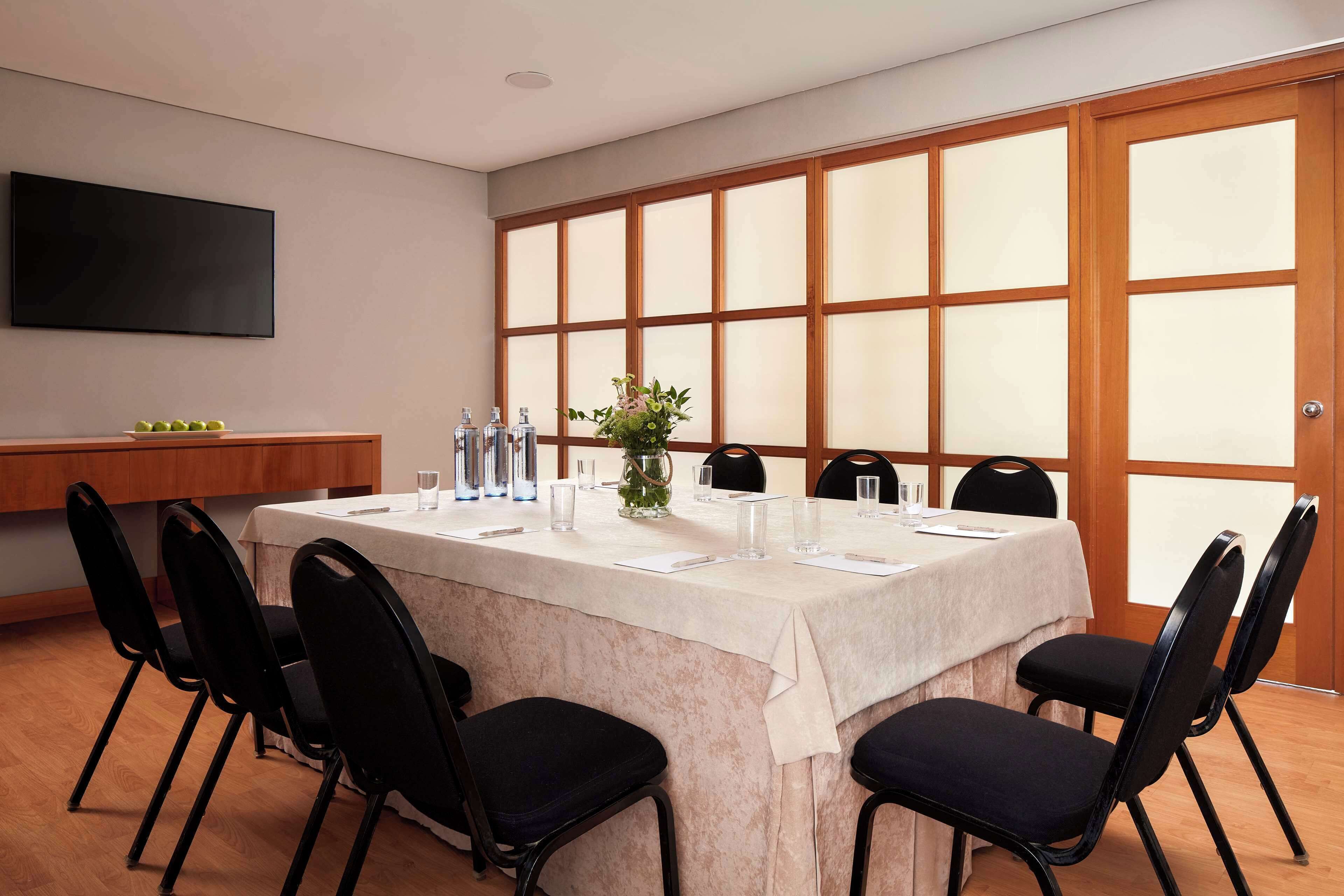 Un salón de conferencias con una larga mesa, sillas negras, un jarrón con flores y una televisión en la pared.
