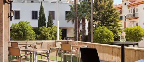 Un patio exterior con mesas y sillas, rodeado de vegetación y edificios al fondo.