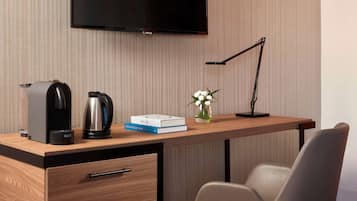 Una habitación de hotel moderna con un escritorio de madera, un televisor de pantalla plana, una cafetera, un hervidor de agua, un libro y un jarrón con flores.