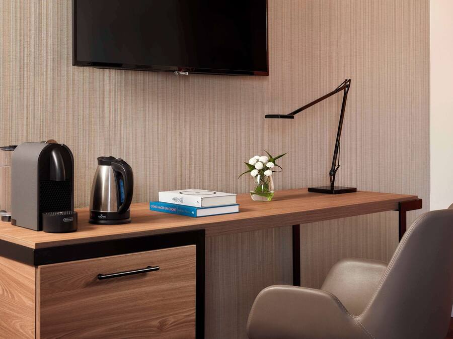 Una habitación de hotel moderna con un escritorio de madera, un televisor de pantalla plana, una cafetera, un hervidor de agua, un libro y un jarrón con flores.