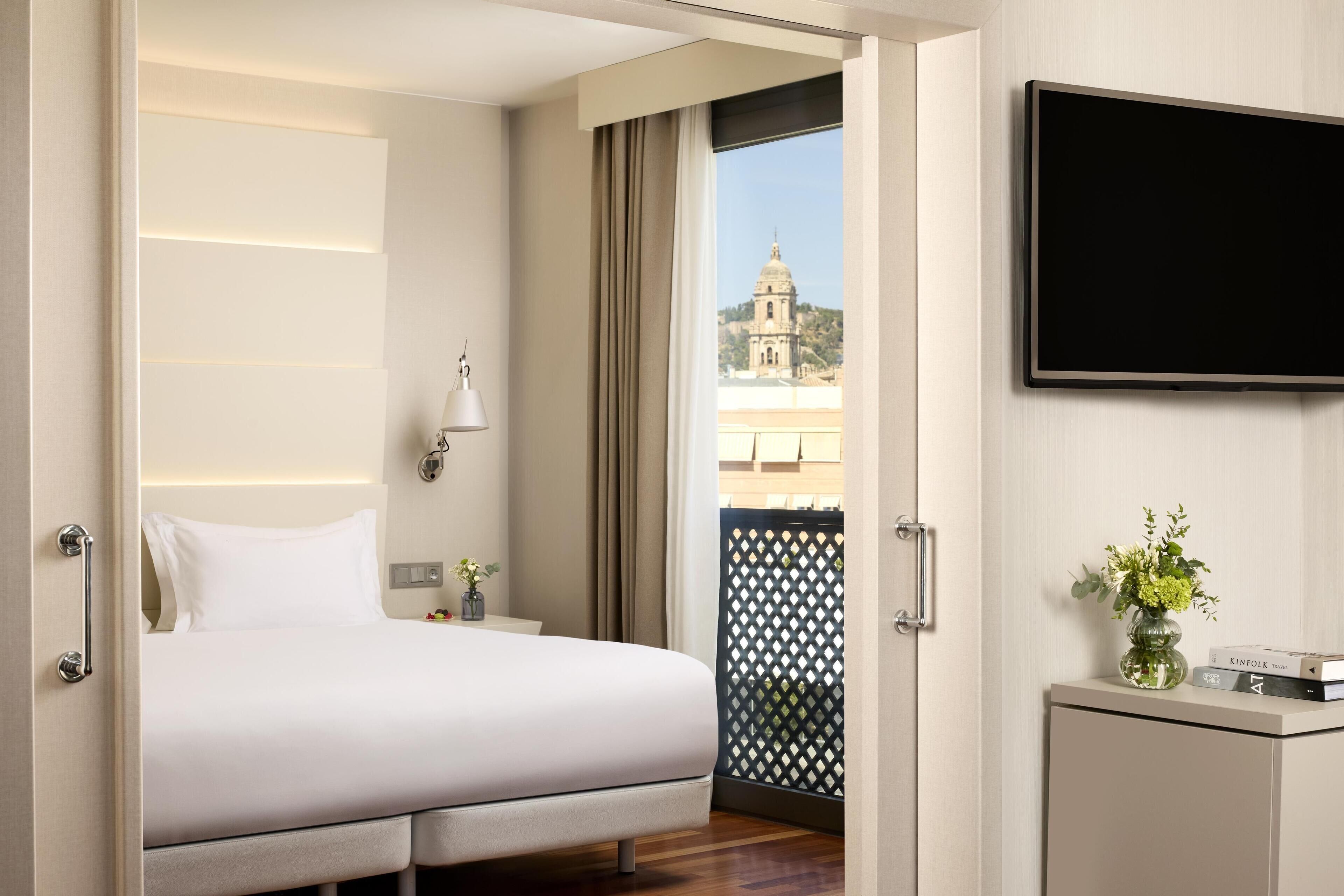 A hotel room with a bed, a TV mounted on the wall, a small table with a vase of flowers, and a view of a building through the window.