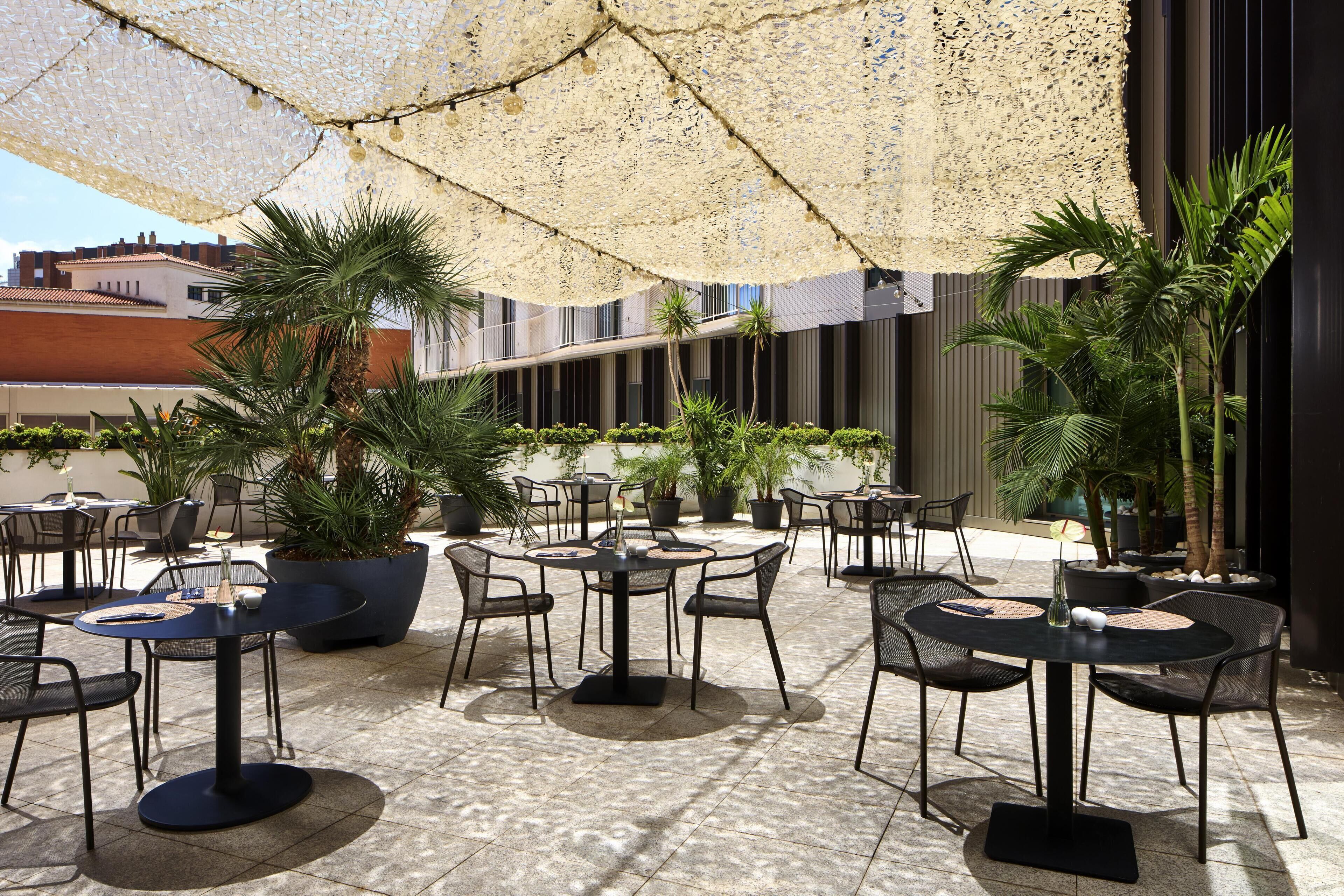 An outdoor patio area with round tables and chairs, surrounded by potted plants and covered by a textured canopy.