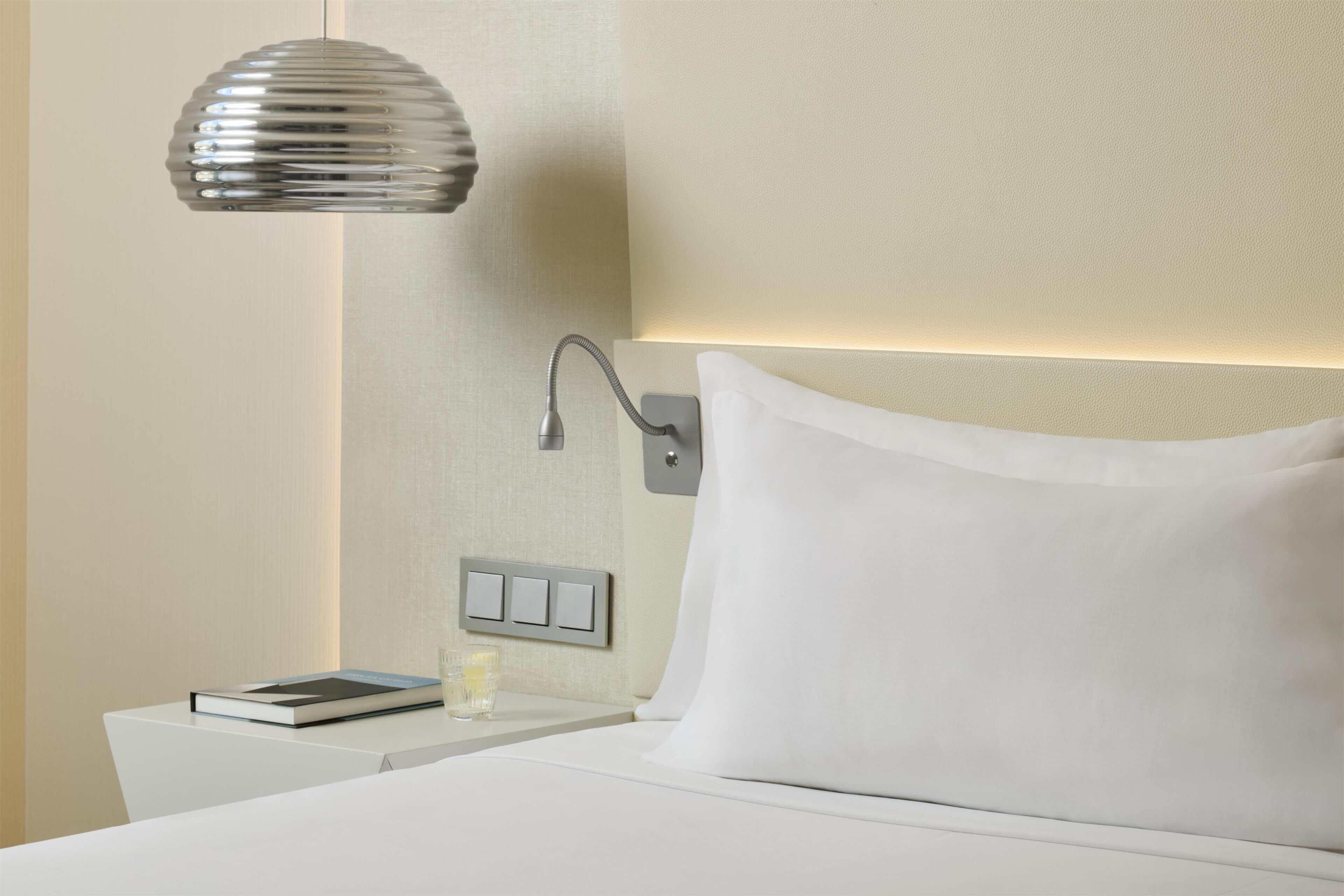 A neatly made bed with white linens, a bedside table with a book and a glass, and a wall-mounted light fixture.