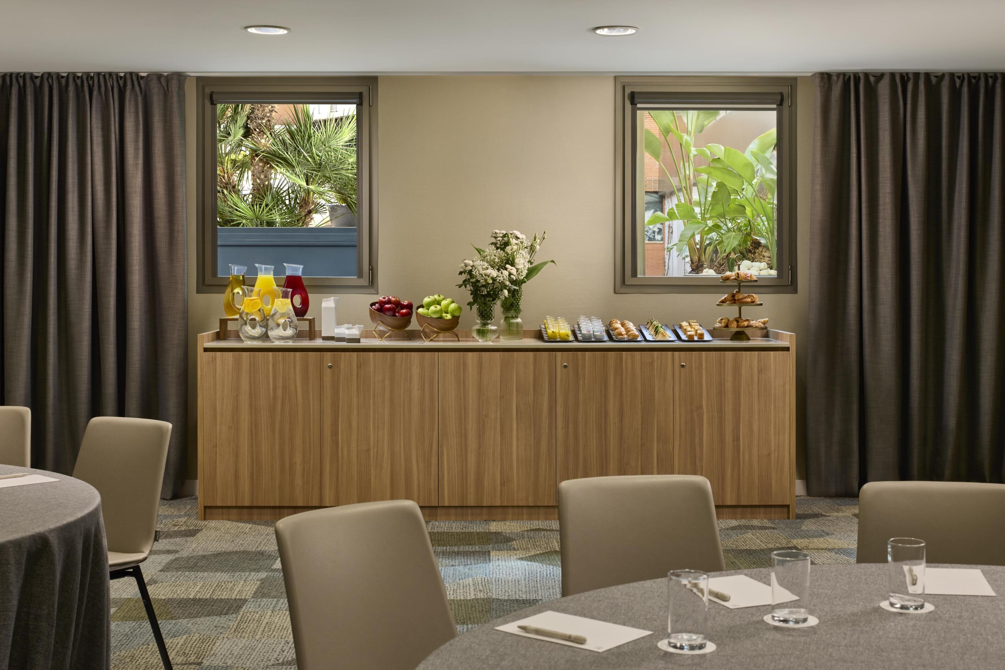 A hotel room with a wooden desk, chairs, a buffet table with food and drinks, and large windows with curtains.