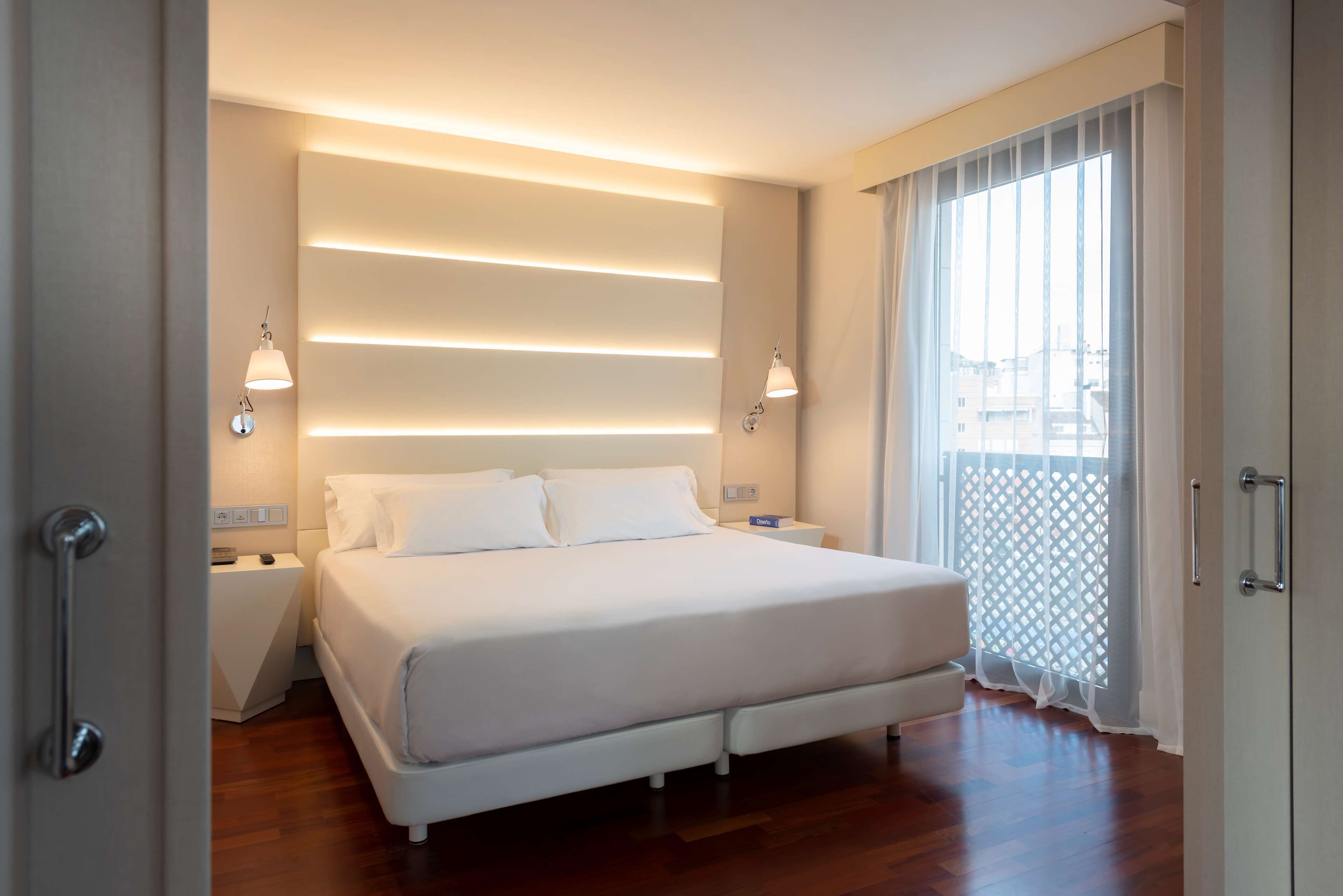 A modern bedroom with a large bed, bedside lamps, and a view through a window with sheer curtains.