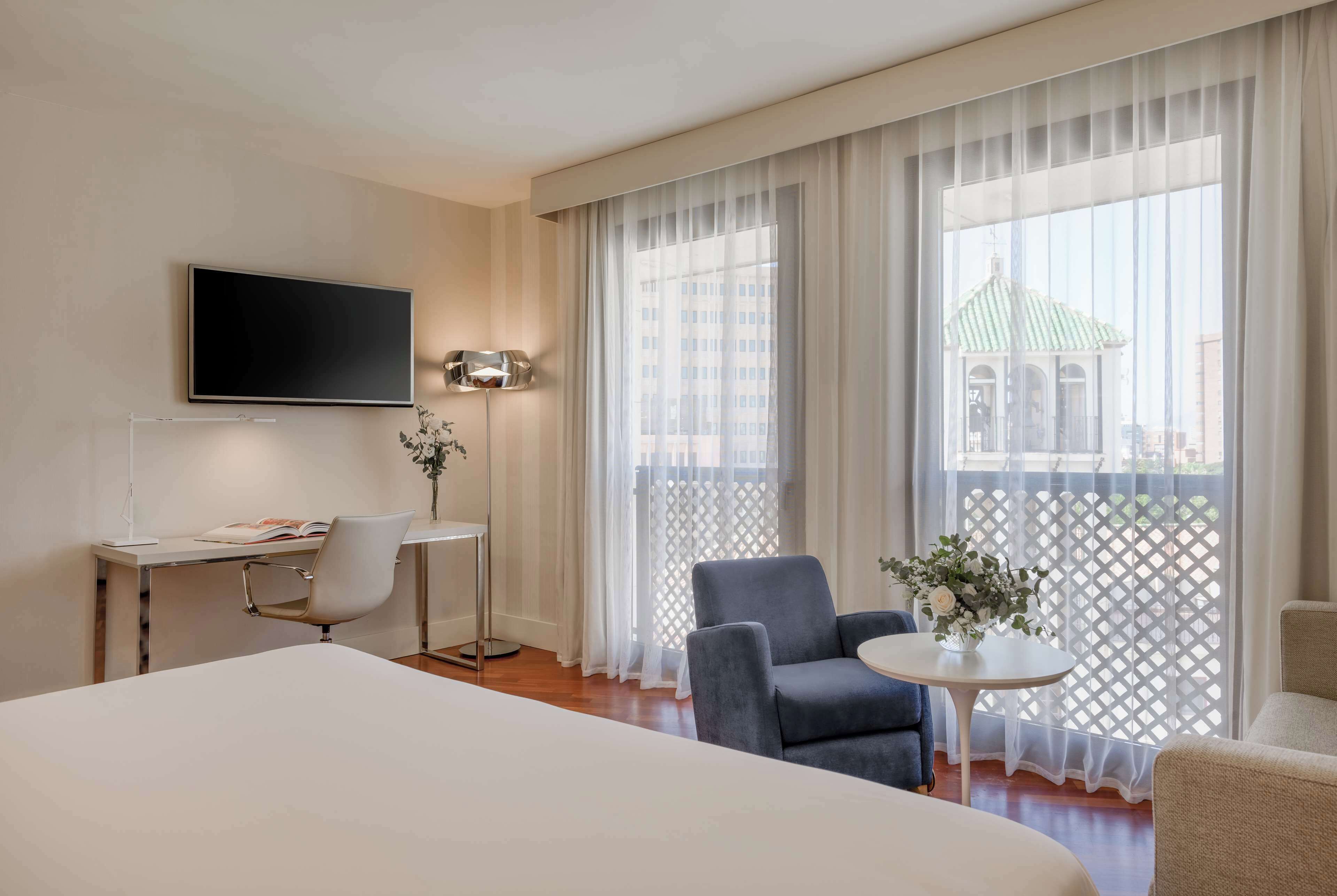 Ein Hotelzimmer mit einem großen Bett, einem Schreibtisch mit Stuhl, einem kleinen Tisch mit Vase und einem Fernseher an der Wand.