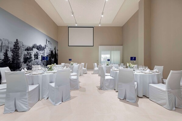 Un salón de conferencias con sillas y mesas blancas, asientos numerados y una pantalla grande en la pared.