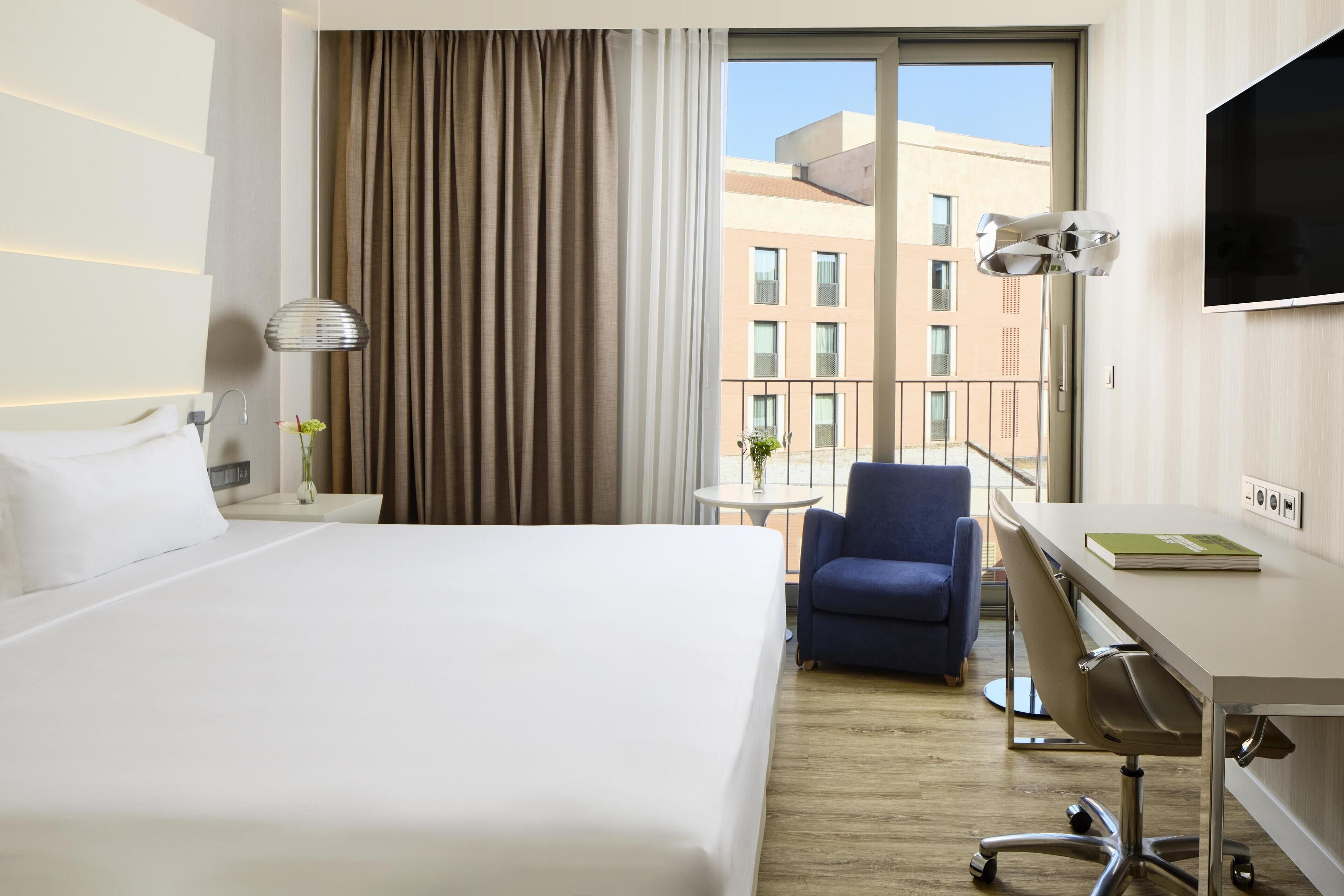 Habitación de hotel con una cama grande, un escritorio con silla, un televisor y vistas a edificios a través de la ventana.