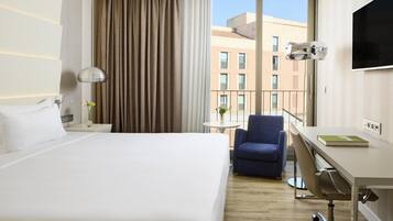Habitación de hotel con una cama grande, un escritorio con silla, un televisor y vistas a edificios a través de la ventana.