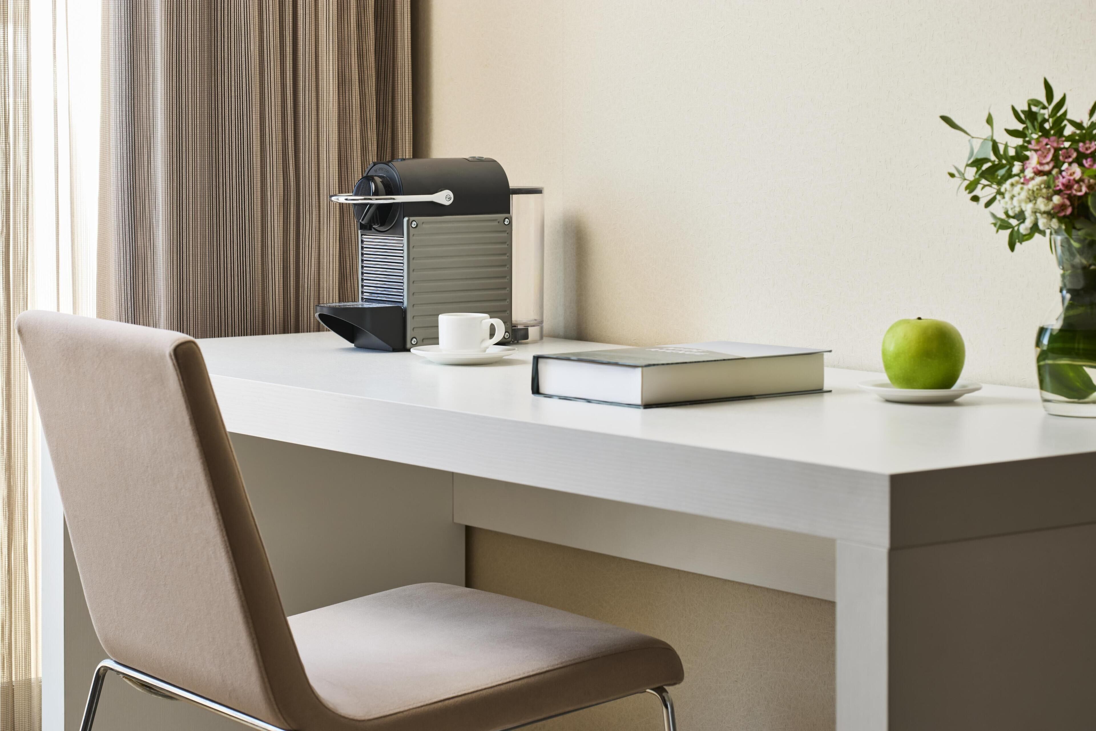 Une chambre d’hôtel moderne avec un bureau blanc, une machine à café, une tasse, un livre, une pomme verte et un vase rempli de fleurs.