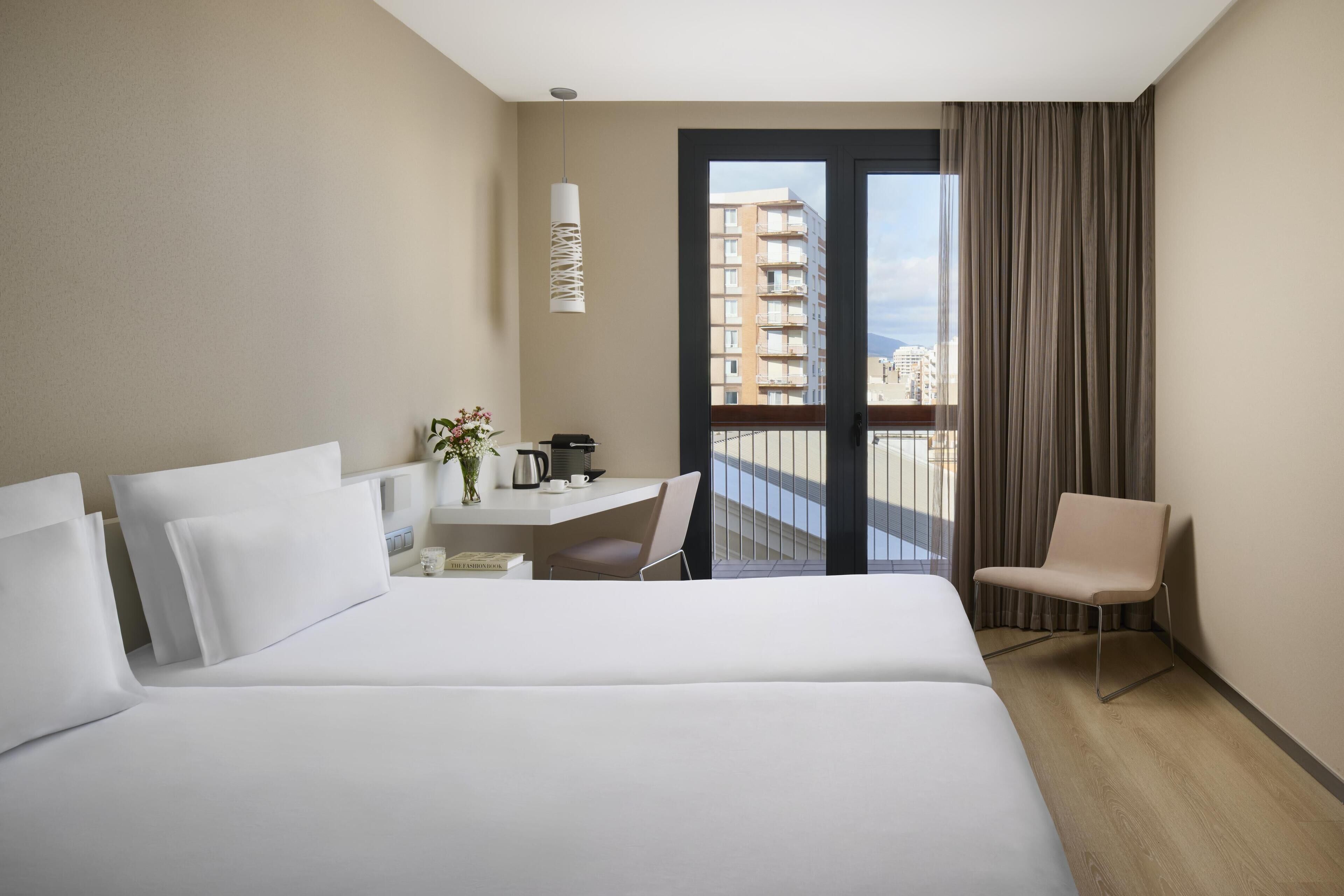 Habitació d'hotel amb un llit gran, una taula de treball amb cadira, una taula petita amb un ram i una paperera, i vista a un paisatge urbà a través d'un balcó amb cortines.