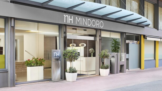 A modern hotel entrance with a glass canopy, large potted plants, and a visible reception desk.