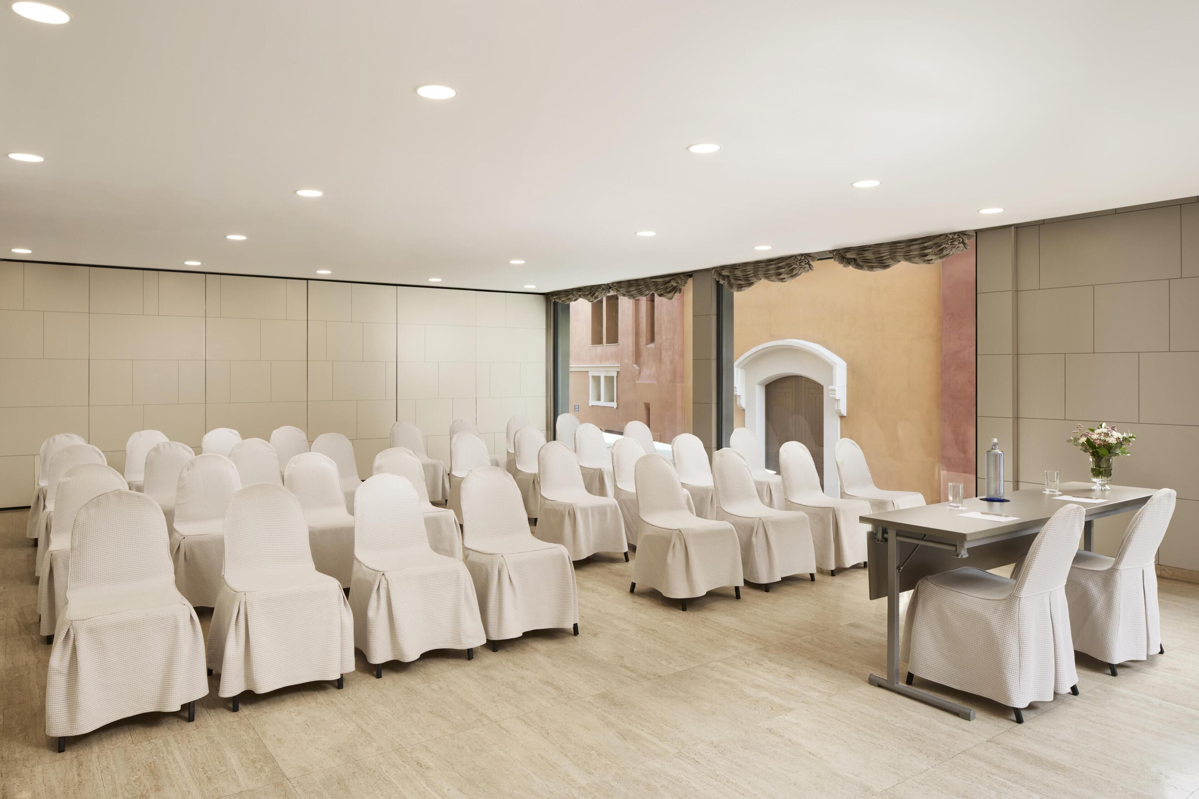Une salle de conférence avec des rangées de chaises blanches et une table entourée de chaises, un plafond blanc et des murs d’une couleur neutre.