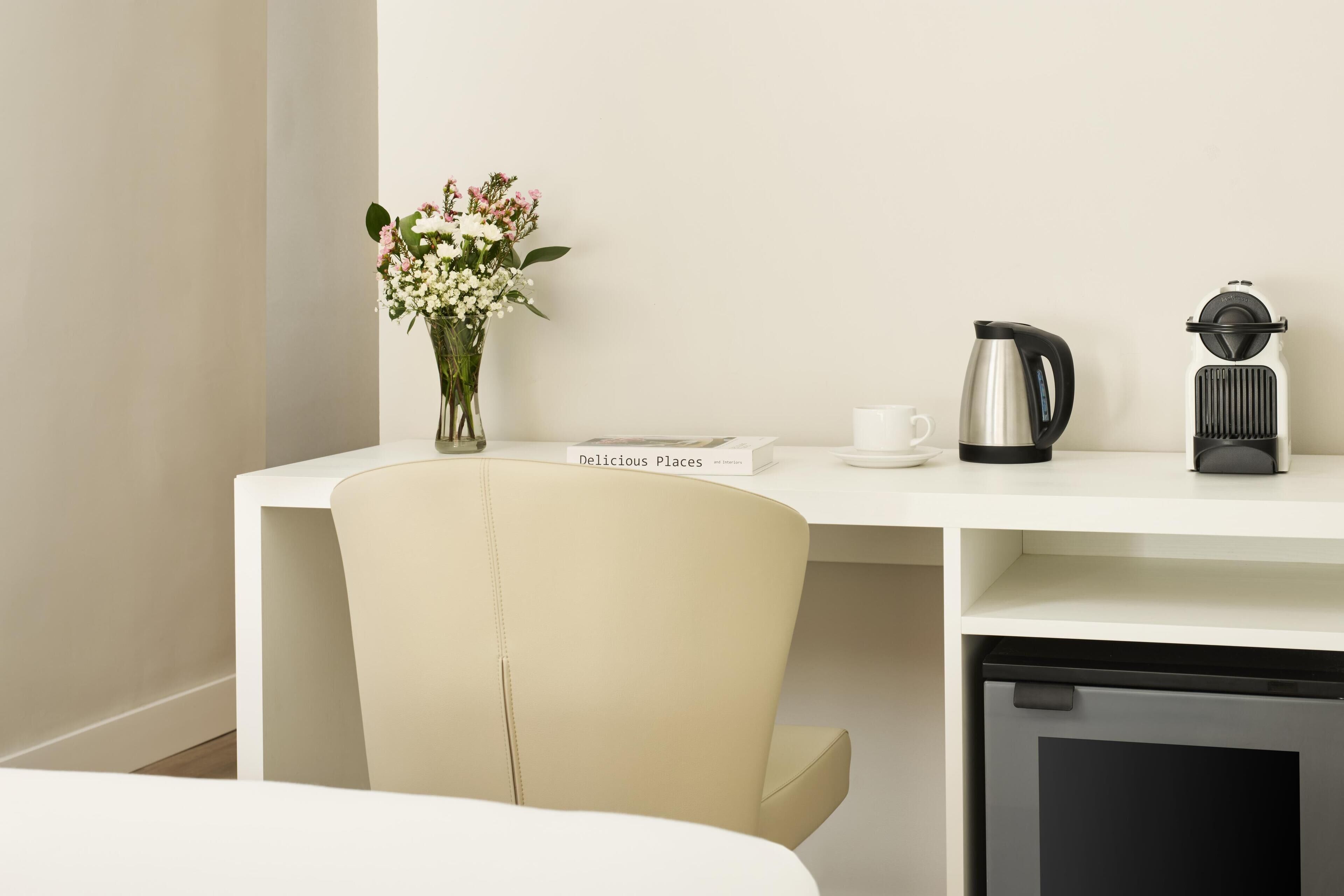 Et moderne, minimalistisk værelse med et hvidt skrivebord, en vase med blomster, en bog med titlen 'Delicious Places', en kaffemaskine og en elkedel.