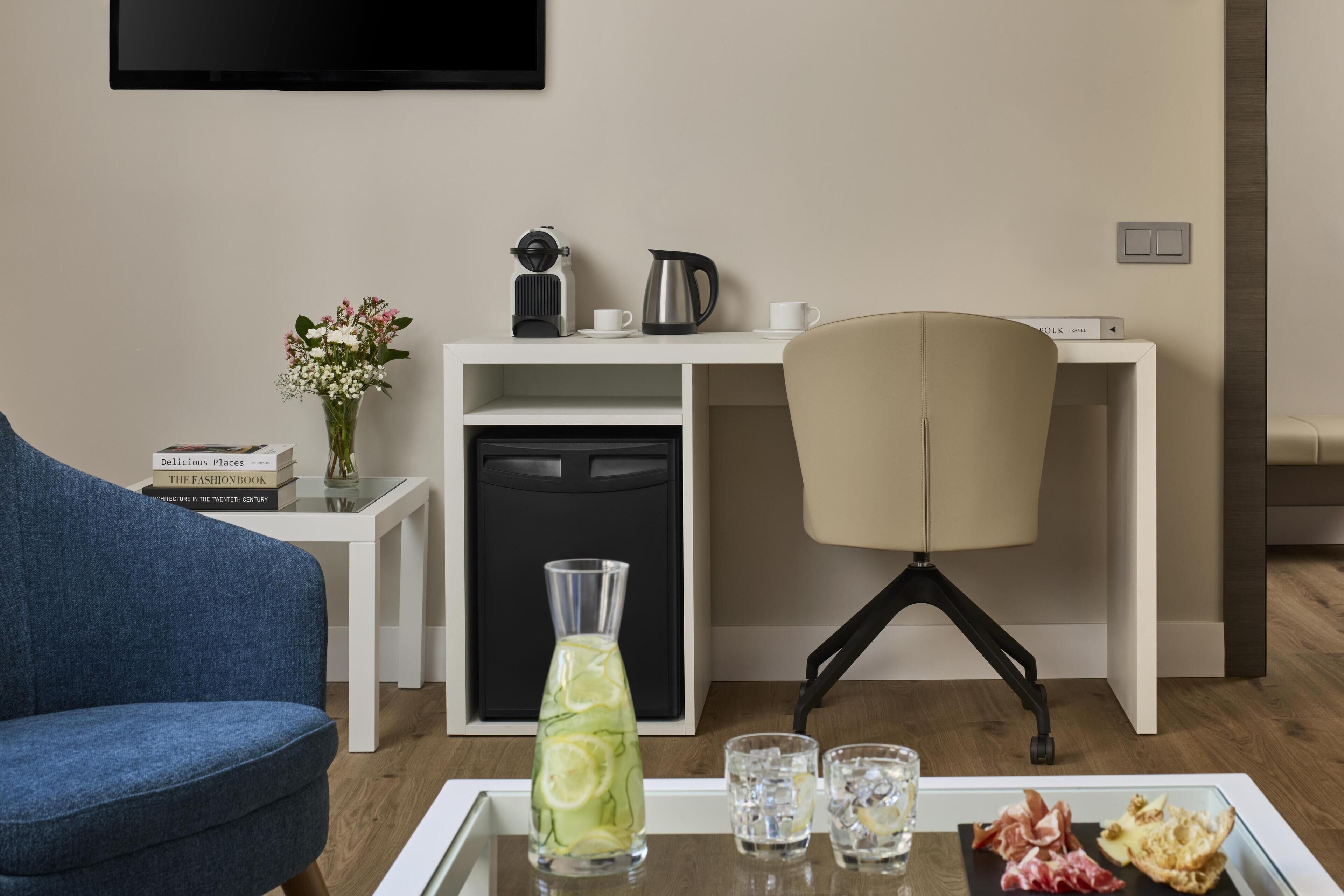 Una sala de estar moderna con un sofá azul, una mesa de centro blanca con bebidas y aperitivos, un sillón beige, un escritorio blanco con un hervidor y un televisor montado en la pared.